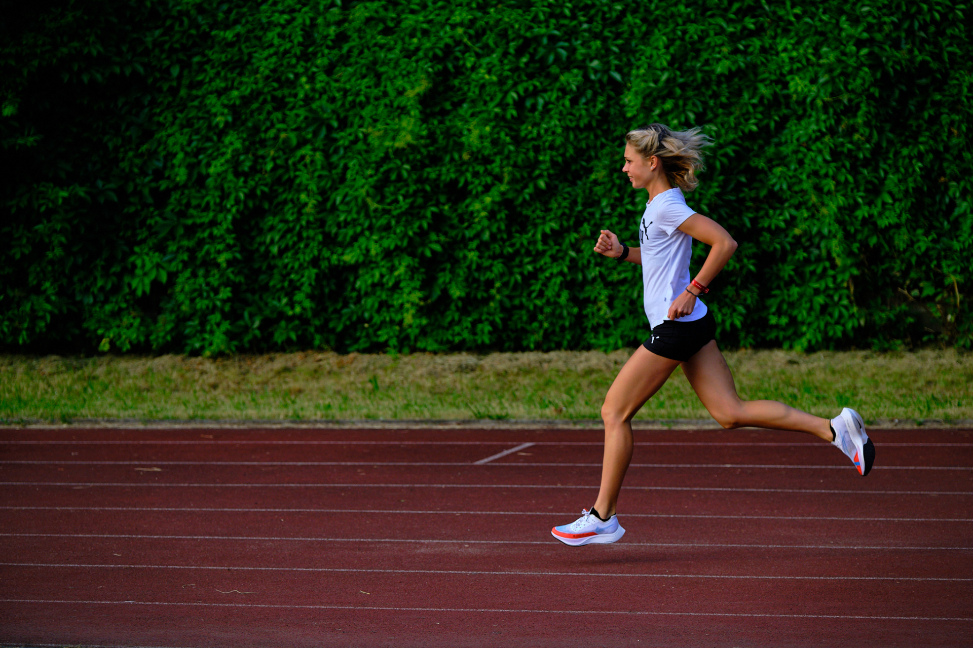 Julianna Nagy fehér atlétatrikóban intervalledzést fut a szabadtéri atlétikai pályán